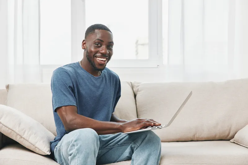 Happy young African American man sitting on a couch, using a laptop and smiling, in a bright and cozy living room with modern decor