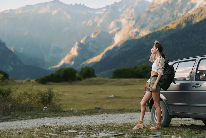 A woman traveler leans against a rugged car in a mountain landscape, backpack beside her. An outdoorsy moment of travel, hiking, nature, and adventure.