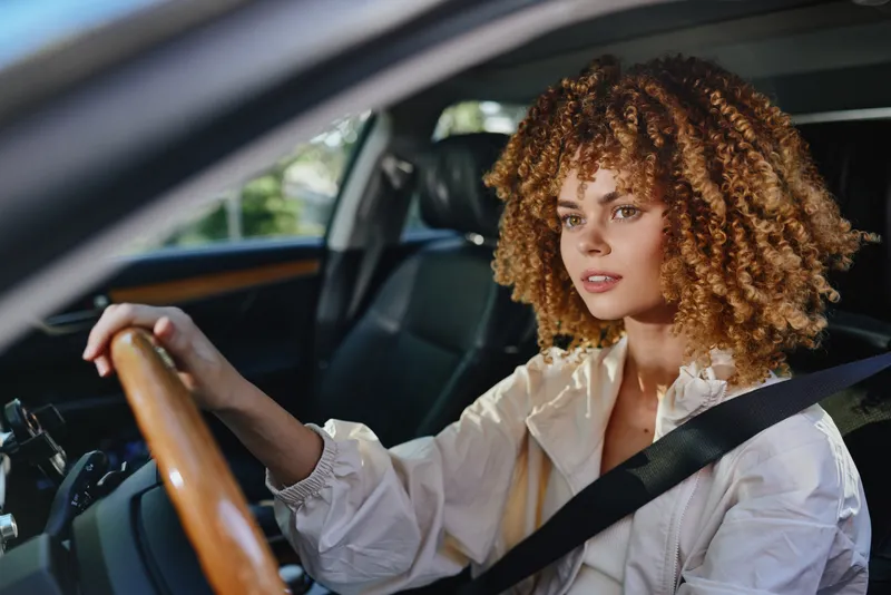 Young woman driving a car with curly hair and focused expression, wearing a seatbelt and light-colored jacket inside a modern vehicle interior.