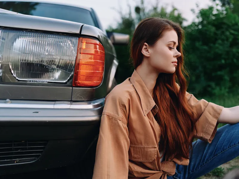 Young woman with long hair in casual attire sitting beside a vintage car, exuding a stylish and relaxed vibe in a natural outdoor setting.