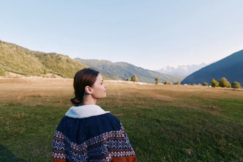 Woman stands in meadow valley looking at mountains and nature landscape outdoors, travel portrait in sweater with serene scenery, grass, sky and panoramic alpine view.