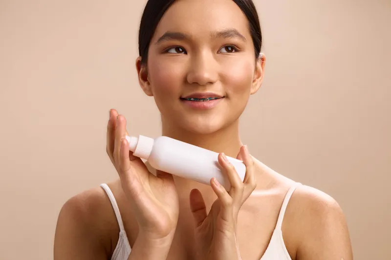 Asian woman applies skincare product in a softly lit room, squeezing lotion from a bottle, focused and calm, conveying a fresh, healthy beauty routine with gentle elegance.