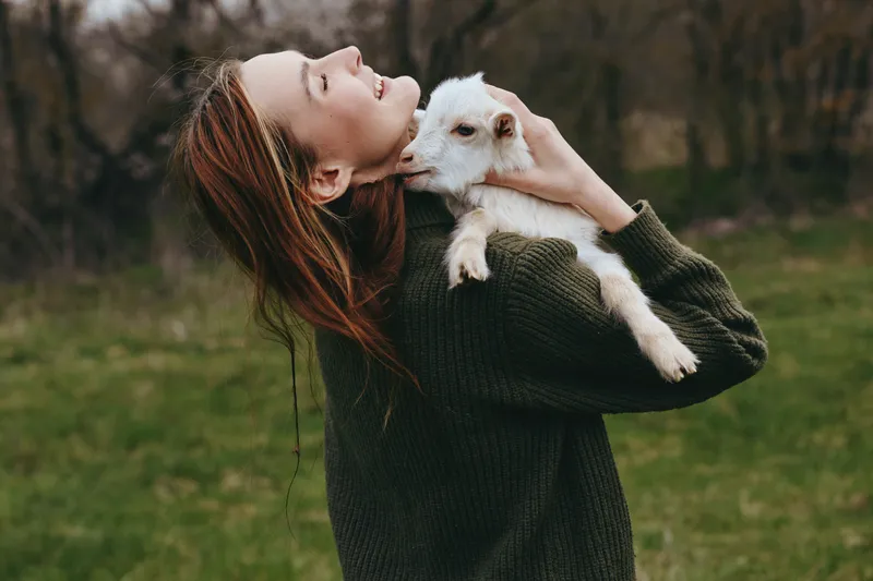 Cheerful woman outdoors, hugging a joyful goat kid, warm sweater, soft breeze, autumn light, relaxed posture, natural colors, meadow background, vibrant greenery, candid moment outdoors