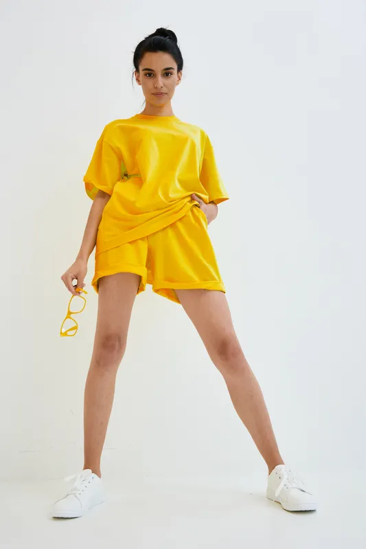 young woman wearing bright yellow casual outfit with white sneakers holding matching sunglasses, standing confidently against white background