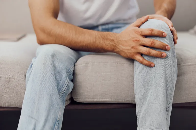 A young man sitting on a couch, gently holding his knee in discomfort. The image captures a moment of vulnerability with a soft color palette and tranquil home setting.