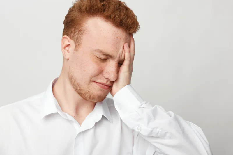 Young redhead man in white shirt feels stressed and overwhelmed, holding his face with closed eyes. Concept of anxiety, frustration, and emotional struggle in everyday life.