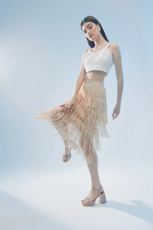 Young woman in a white crop top and beige fringed skirt poses gracefully on a light background. Concept of woman beauty fashion, elegance, and modern style in studio lighting.