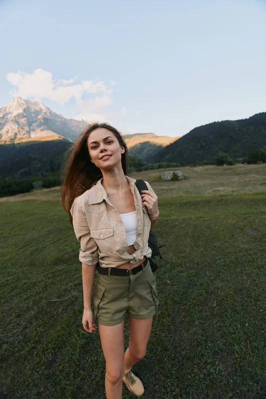 Young woman trekking in an open meadow with distant mountains, wearing a beige jacket and shorts. Natural light highlights a casual lifestyle outdoor moment of exploration.