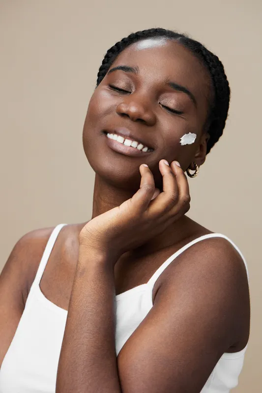 Smiling African woman applying skincare cream on her face with a warm background, capturing the essence of self care and beauty in a minimalist style