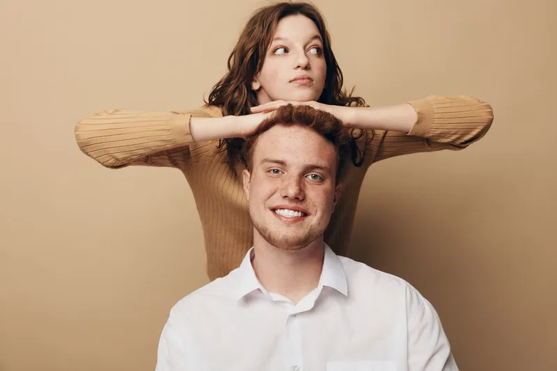 Smiling young man in white shirt with curly woman in brown sweater resting hands on his head. Studio portrait on beige background. Concept of friendship, casual style, and positive mood.