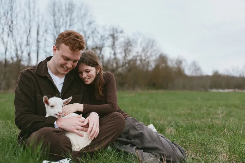 A couple sits on a grassy field, embracing a small goat kid in their lap, smiling softly. The moment captures gentle rural lifestyle, care for animals, and a peaceful connection in nature.