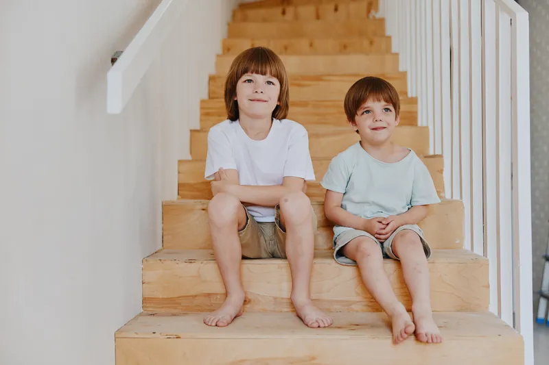 Two happy boys sitting on wooden stairs, enjoying their day with casual clothing and barefooted, radiating a joyful atmosphere and carefree spirit.