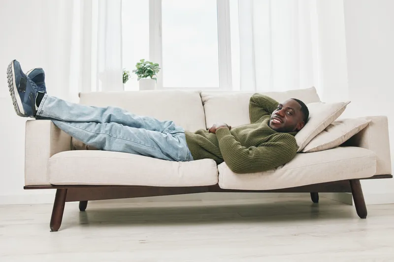 Happy man relaxing on a modern couch in a light filled room, wearing casual attire and enjoying a peaceful moment at home, embodying comfort and tranquility