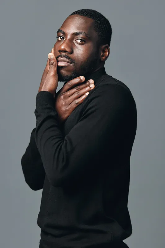 Creative conceptual portrait of African man in black sweater posing with hands near face against gray background. Stylish visual art with focus on expression and skin texture.