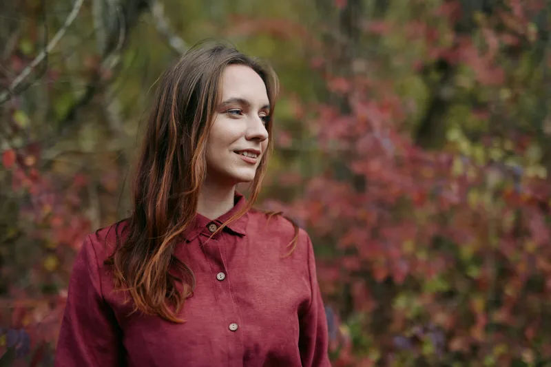 A candid portrait of a young woman standing among autumn foliage, her calm gaze and natural expression conveying authenticity and honest presence.