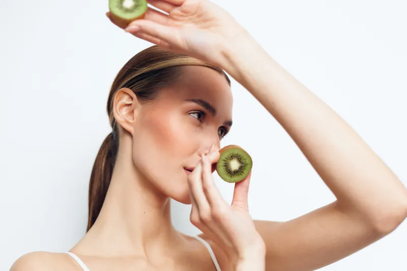 Young woman holds kiwi fruit slices on her fingertips, touching her face and looking to the side, concept of fresh skincare routine and natural beauty care, white studio background