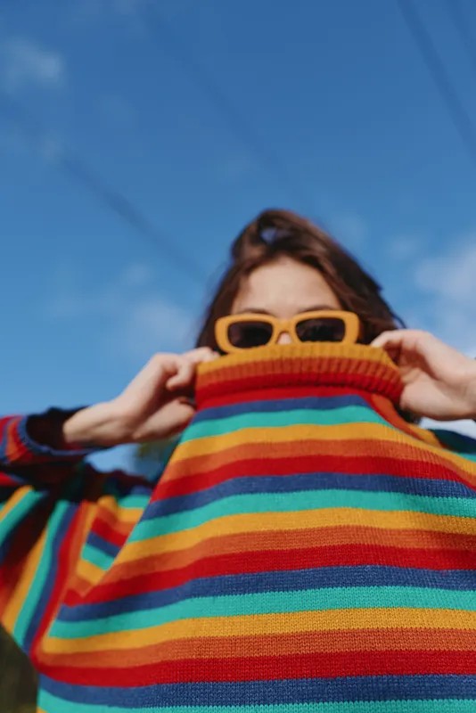 Sweater stripes sunglasses portrait woman colorful fashion against blue sky, playful closeup of person pulling turtleneck over face in outdoor casual style and bright knitwear mood.