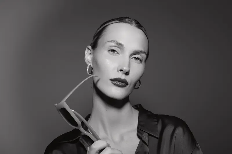 A fair skinned Caucasian woman with slicked back hair holds sunglasses near her chin in a monochrome studio portrait. Concept of fashion, elegance, and beauty.