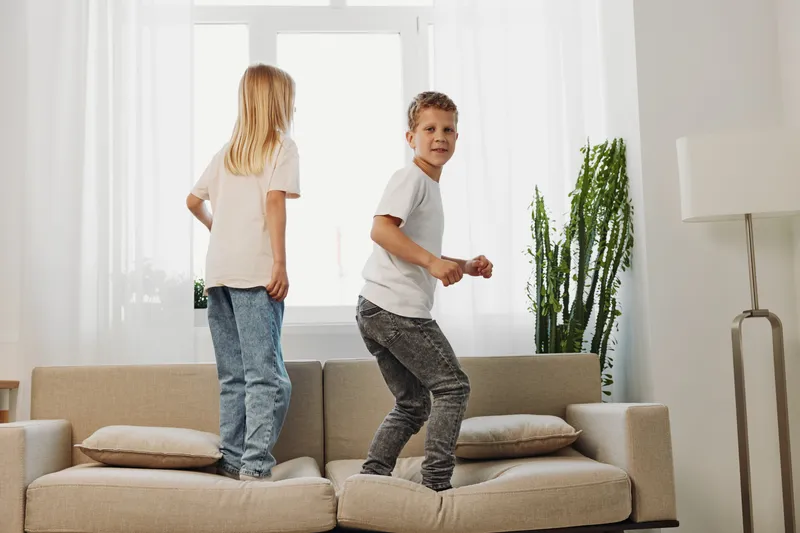 Two playful children jumping on a beige sofa in a bright living room with large windows and green plants. Concept for childhood fun, energy, and home activity.