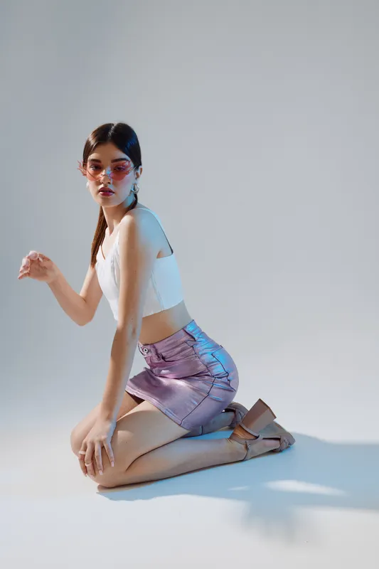 Young woman beauty fashion model kneels on the floor wearing a white crop top and purple skirt with heels. Studio shot with soft lighting on a plain background. Concept of style and elegance.
