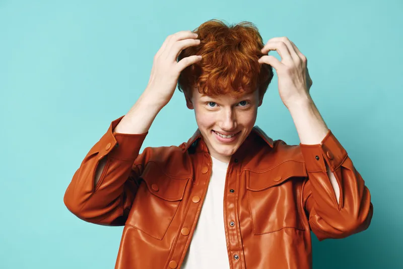 Redhead teenage boy smiling and touching hair wearing brown leather jacket and white shirt against blue background with playful and confident expression.