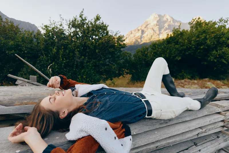 Woman relaxation in nature lying outdoors on a wooden fence with mountains in the background, countryside fashion portrait wearing boots, denim vest and shearling jacket for peaceful escape.