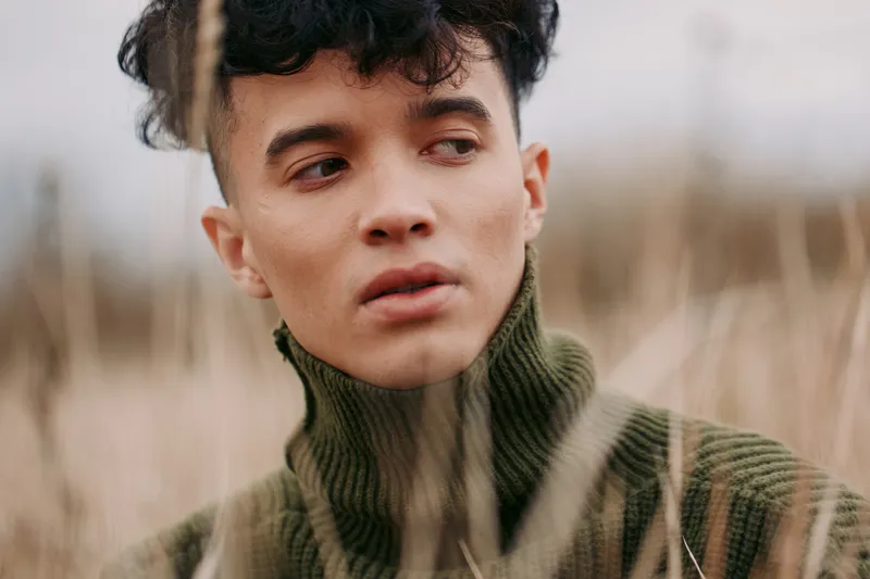 A candid portrait of a young man in a green knit turtleneck, conveying authenticity and credibility with soft focus and natural light in a misty field.