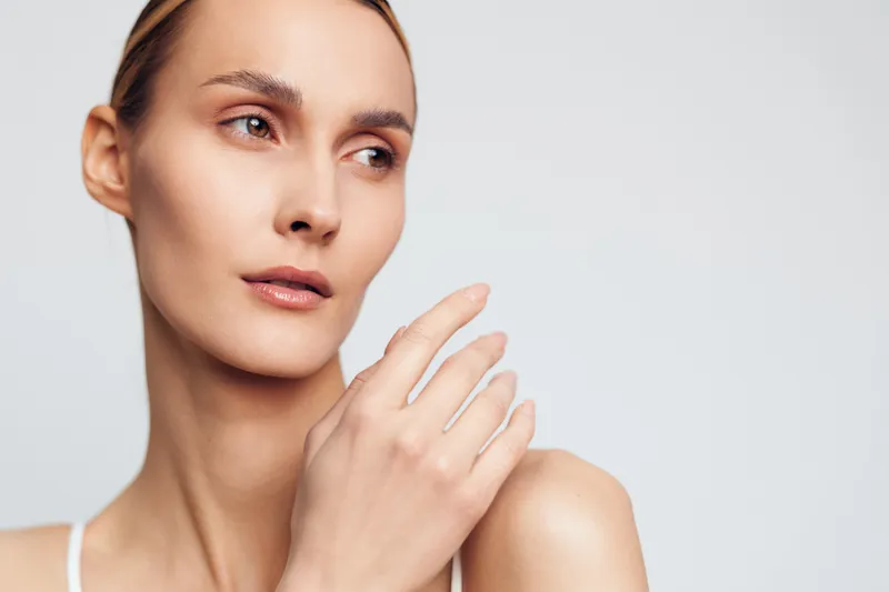 Young woman with natural skin looks away thoughtfully with hand near face isolated on white background, showing clean complexion and soft delicate features in studio lighting.