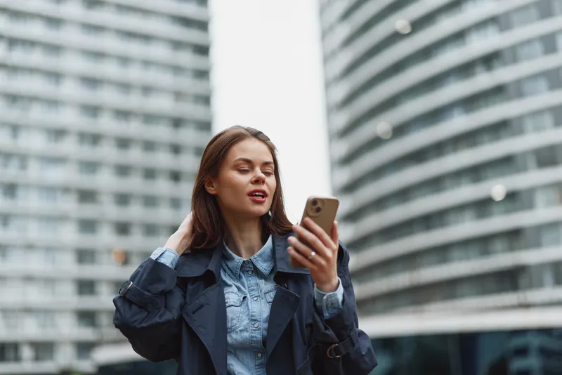 Young woman in a denim shirt and navy coat checks her smartphone in an urban plaza with glass buildings behind.