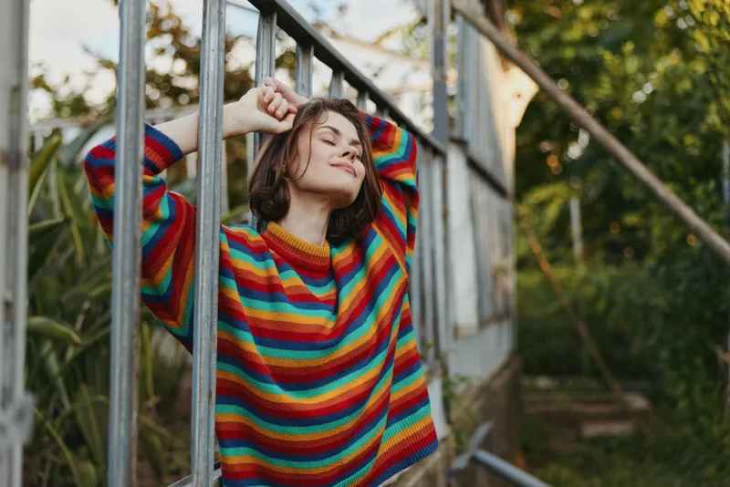 Woman in rainbow sweater smiling and relaxed outdoors, portrait leaning on metal railing among greenery. Casual fashion, cozy knit and warm natural light capturing a peaceful moment.