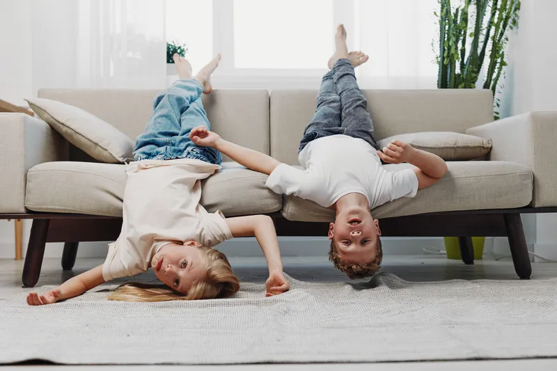 Two playful children hang upside down from a beige sofa in a bright living room, enjoying fun and carefree moments of childhood indoors.