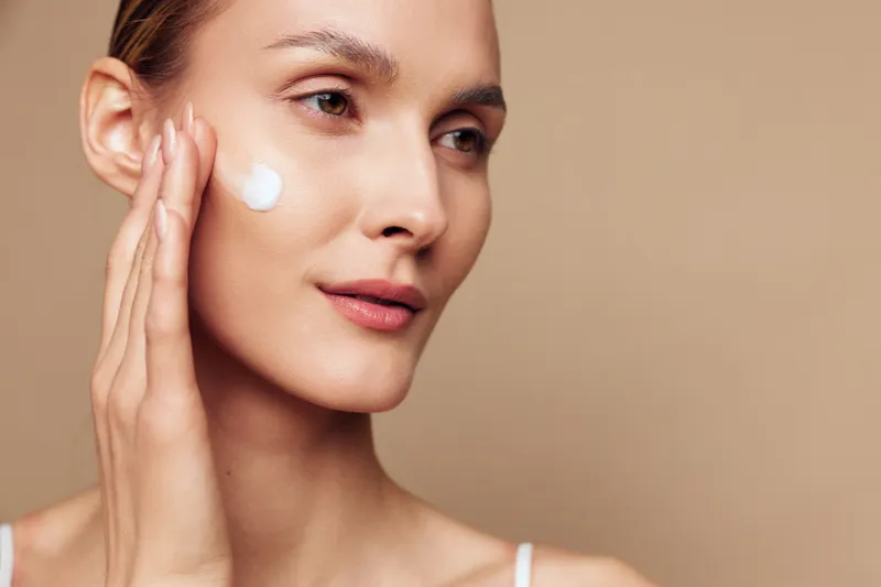 Young woman applies moisturizing cream on face with gentle touch looking away with calm expression isolated on beige background skin care concept natural beauty closeup portrait.