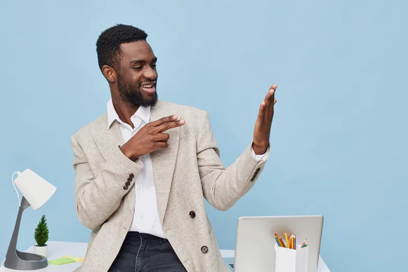 Confident young man with dark skin and short curly hair, dressed in beige blazer and white shirt, looking at smartphone and smiling, sitting in modern office with blue background. People lifestyle con