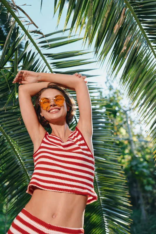 A cheerful woman in a red striped crop top and round sunglasses smiles while posing outdoors beneath palm leaves, radiating sunny energy and carefree summer vibes.