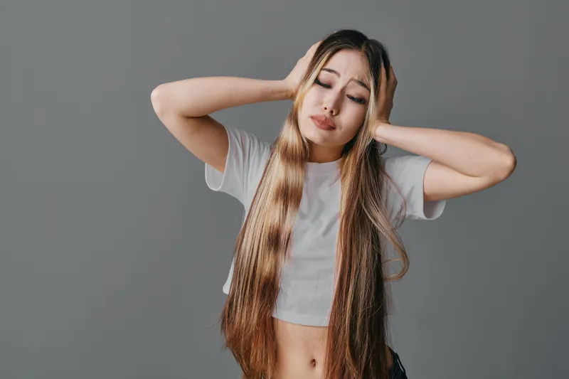 Young woman standing with long hair and closed eyes holding head in pain on isolated background. Female expressing stress, headache or discomfort with casual white shirt.