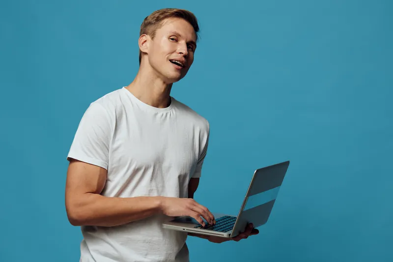 smiling man holding laptop wear white t-shirt look camera standing isolated on blue background work in metaverse digital nomad concept