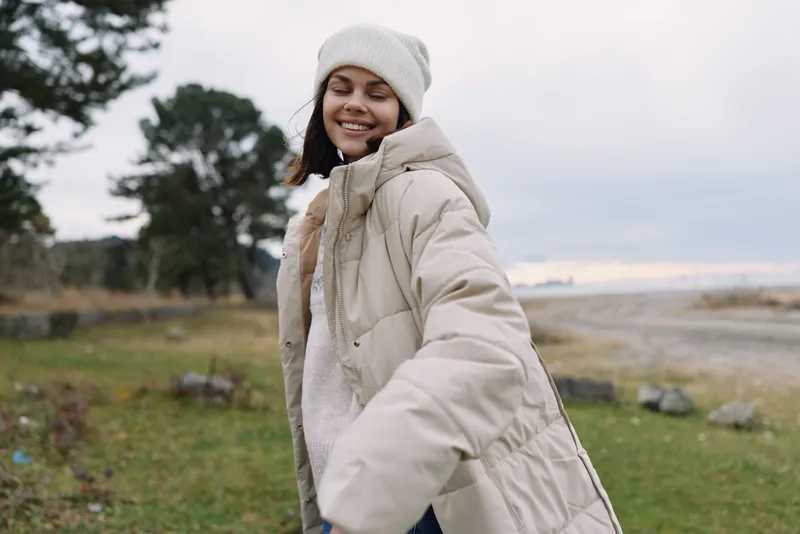 Woman in a warm beige coat smiles outdoors in a windy countryside, wearing a knit hat and cozy sweater as soft clouds drift over a grassy field and trees.