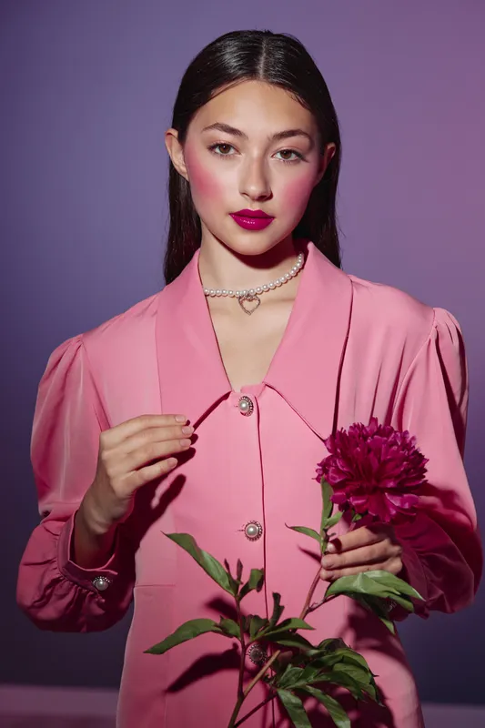Young woman in pink stylish outfit holds a burgundy flower while wearing bracelet and dark long hair. Vibrant makeup emphasizes natural beauty. Creative fashion portrait concept.