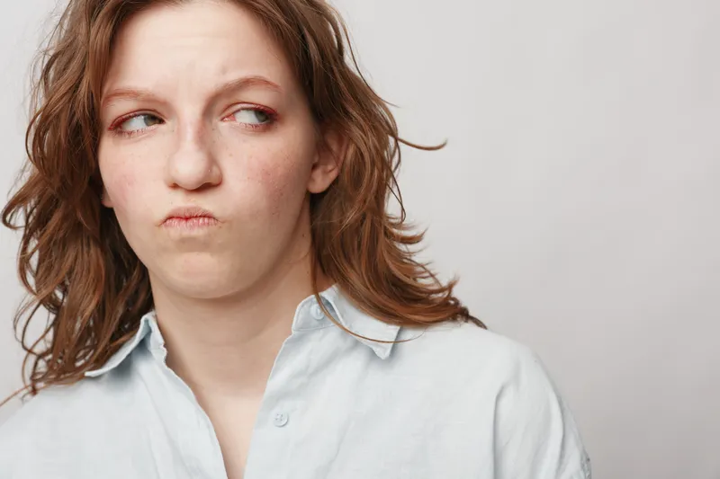 Young woman with curly hair looks sideways with a skeptical expression wearing a light shirt. Neutral background highlights her doubtful and uncertain mood concept.