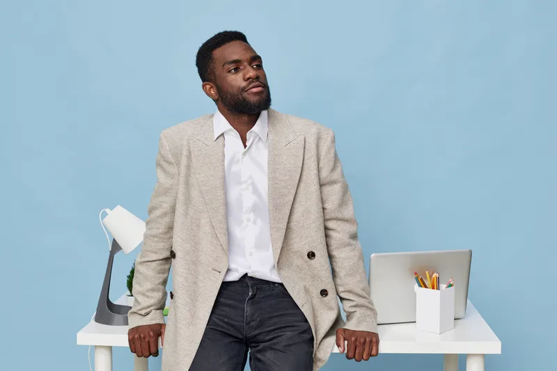 Confident man in smart casual attire leaning on office desk against solid light blue background, thoughtful expression, modern workspace, professional atmosphere, business casual style, minimalistic c