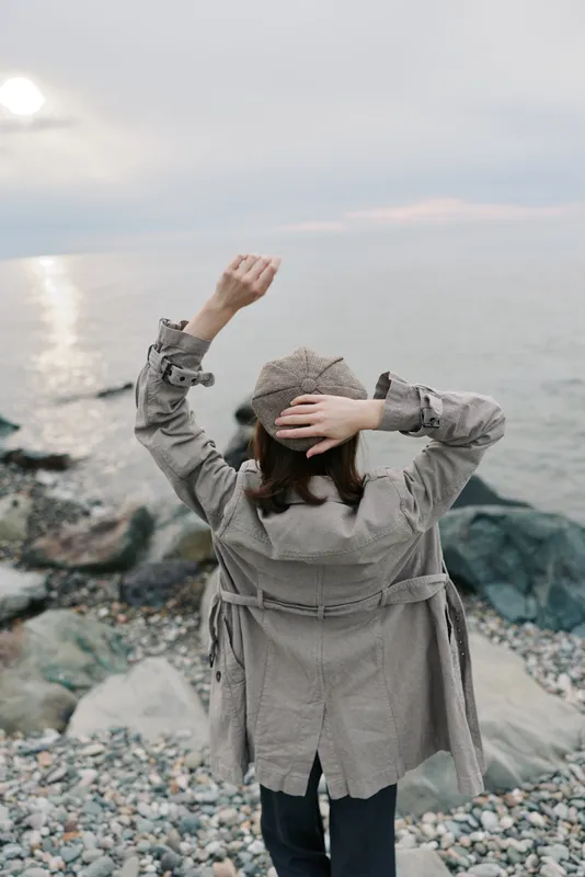 Woman in gray coat and hat stands on rocky shore facing calm sea and cloudy sky. Concept of solitude, nature, and peaceful coastal landscape during overcast weather.