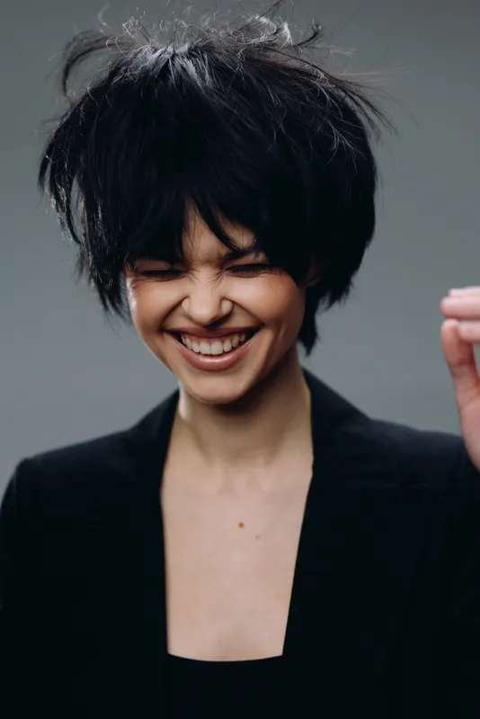 Smiling woman with messy black hair wearing black jacket, expressing joy and freedom with closed eyes, isolated on gray background.