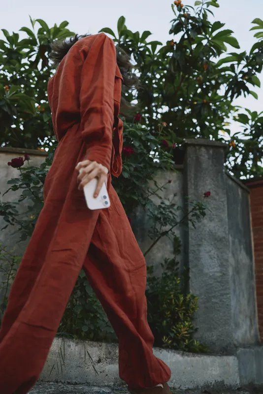 A person in a red jumpsuit walks along a sidewalk, phone in hand, captured from a low angle. A candid woman lifestyle moment highlighting movement, casual fashion, and everyday commute
