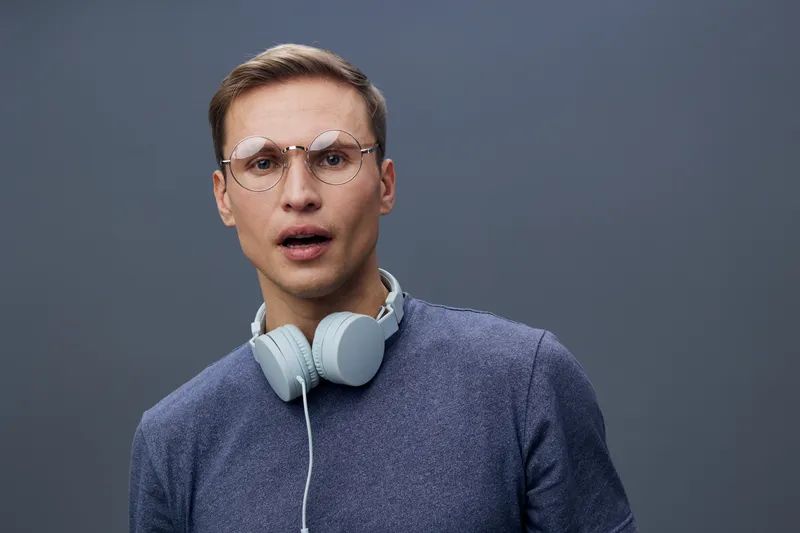 Confused young man wearing glasses and gray sweater with headphones around neck looking surprised against plain gray background studio portrait. People lifestyle concept