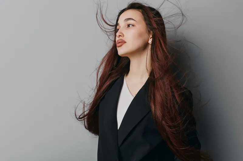 Young woman with long hair wearing black blazer and white shirt stands against gray background looking sideways with wind blowing through her hair for fashion portrait concept.