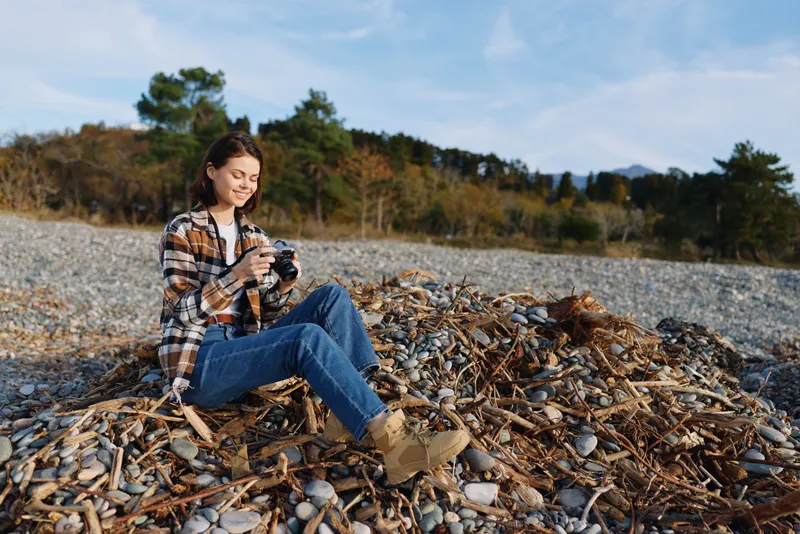 Woman with camera sits on a pebble beach, wearing a plaid shirt and jeans, capturing outdoor scenery with trees and distant hills under a blue sky.