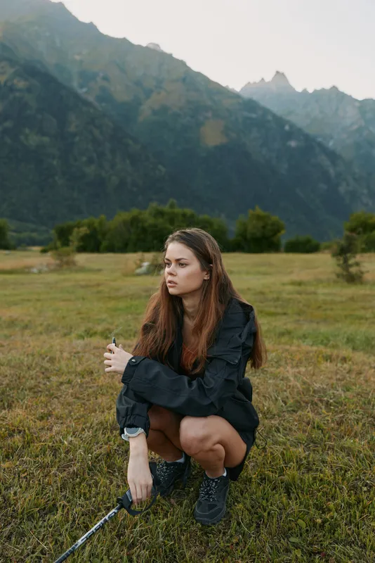 A woman crouches in a grassy meadow, mountains rising in the background, wearing a dark jacket. She gazes sideways with a thoughtful, calm expression under soft light.