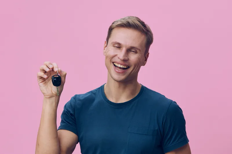 Smiling young man with short brown hair wearing a blue t-shirt holding a car key on a pink background studio portrait. People lifestyle concept