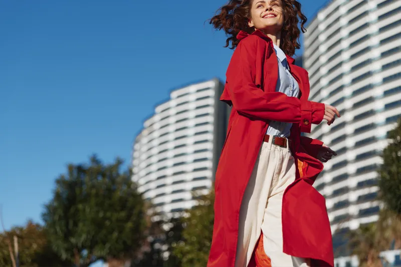 A stylish woman in a redcoat walks outdoors in a sunlit cityscape, exuding confident charm as modern buildings rise behind her and a soft breeze lifts her hair.
