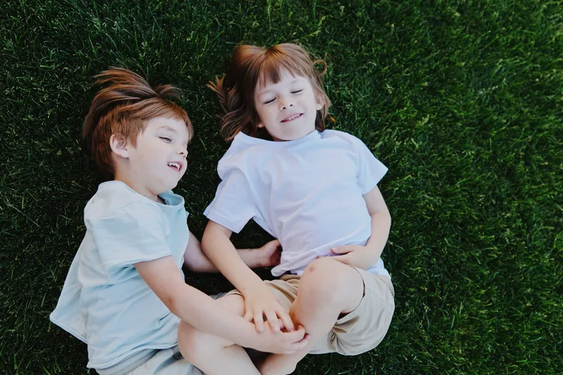Two happy children lying on green grass, enjoying a sunny day. Their joyful expressions capture the essence of childhood and carefree moments outdoors.